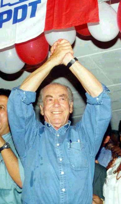 Leonel Brizola com as mãos erguidas sob uma faixa do PDT, com balões vermelhos e brancos